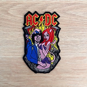 Könnte beinhalten: Ein gestickter Bügel-Patch mit dem AC/DC-Logo und einem Cartoon-Bild eines Teufels und eines Mannes im Anzug. Der Teufel ist rosa mit roten Haaren und Hörnern, und der Mann trägt einen blauen Anzug und einen Hut.