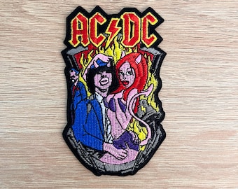 Op de afbeelding: Geborduurde patch met het AC/DC-logo in rood en geel, en een cartoonillustratie van twee figuren. Eén figuur draagt een blauw pak, de andere is een roze duivelachtig personage met rood haar. De patch heeft een zwarte rand.