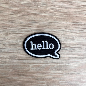 Könnte beinhalten: Schwarzer und weißer gestickter Aufnäher in Form einer Sprechblase mit dem Wort "hello" in weißen Buchstaben.