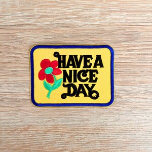 May include: Embroidered patch with a yellow background and a blue border. The patch features a red flower with a green stem and the phrase "HAVE A NICE DAY" in black lettering. A small smiling face is at the bottom.