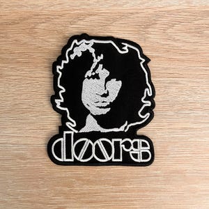 Puede incluir: Parche bordado en blanco y negro con un retrato de una persona con pelo largo y la palabra "doors" en una fuente estilizada. El parche es rectangular con fondo negro y costuras blancas.