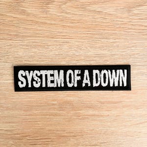 Könnte beinhalten: Ein rechteckiger schwarzer Aufnäher mit dem Text "SYSTEM OF A DOWN" in weißer, verwitterter Schrift. Der Aufnäher liegt auf einer hellbraunen Holzoberfläche, wodurch ein Kontrast zwischen dem dunklen Aufnäher und dem helleren Hintergrund entsteht.