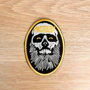Könnte beinhalten: Ovaler, gestickter Aufnäher mit einem Totenkopf mit weißem Bart und dem Wort "MASTODON" in Gelb. Der Totenkopf befindet sich auf einem schwarzen Hintergrund mit gelbem Rand. Für Kleidung oder Accessoires.