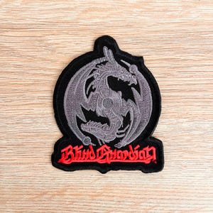 May include: Black and grey embroidered patch featuring a silver dragon design and the text "Blind Guardian" in red.