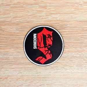 Puede incluir: Un parche circular blanco y negro con una letra 'S' estilizada roja en el centro. La palabra "SHINEDOWN" está escrita verticalmente en blanco en el lado izquierdo del parche.