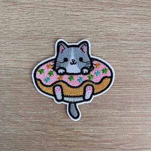 May include: An embroidered patch featuring a gray cat floating in a pink donut. The donut has sprinkles in various colors and a brown outline. The cat has a smiling face and is surrounded by a white border.