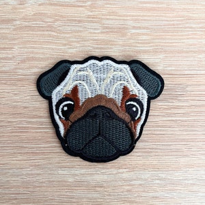 May include: Embroidered patch of a pug dog's face. The pug has brown fur, a black nose, and white fur around its eyes.