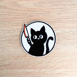 Puede incluir: Parche bordado con un gato negro sosteniendo un cuchillo con una hoja roja y naranja. El gato está dentro de un círculo blanco con un contorno negro. El gato tiene ojos grandes.