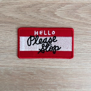 Puede incluir: Un parche rectangular bordado en rojo y blanco con las palabras "HELLO Please Stop" en escritura negra. El parche tiene un borde rojo y un centro blanco.