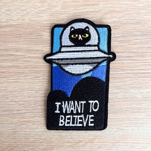 Może przedstawiać: Naszywka haftowana przedstawiająca czarnego kota w latającym spodku na tle niebieskiego i białego nieba. Tekst "I WANT TO BELIEVE" jest napisany na czarnym tle białym kolorem.