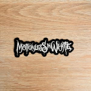 May include: A black and white embroidered patch featuring the words "Motionless In White" in a gothic style font. The patch has a black border and is on a light brown wooden surface.