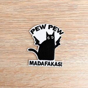 Puede incluir: Gato negro sosteniendo dos pistolas con el texto "PEW PEW MADAFAKAS!" en letras blancas sobre un fondo blanco.