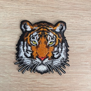 May include: Embroidered tiger head patch. The patch has a black border, with orange, white, and black stripes. The tiger has yellow eyes and a red nose. Suitable for application on clothing or accessories.