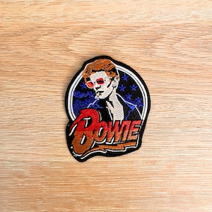 May include: An embroidered patch featuring a stylized portrait of David Bowie with orange hair and red sunglasses. The design includes a dark blue background with small stars inside a circular border, and the word Bowie in large red lettering.