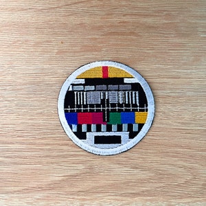 Op de afbeelding: Een ronde geborduurde patch met een zwart-wit televisie testpatroon. Het patroon heeft een gele, rode, groene, blauwe en witte kleurstelling.