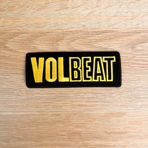 Könnte beinhalten: Schwarzer rechteckiger Aufnäher mit dem Wort "VOLBEAT" in gelben Blockbuchstaben gestickt. Der Aufnäher liegt auf einer hellbraunen Holzoberfläche.