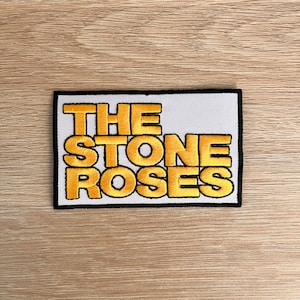 May include: Embroidered patch featuring the text "THE STONE ROSES" in bold, yellow letters with a black outline, set against a white rectangular background. The patch has a black border and is likely for clothing or accessories.