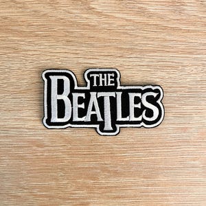 Może przedstawiać: Czarna i biała naszywka z napisem "THE BEATLES" w odważnej, stylizowanej czcionce. Naszywka ma białą obwódkę i prawdopodobnie jest przeznaczona do przyszywania lub naprasowywania na tkaninę. Naszywka leży na jasnobrązowej drewnianej powierzchni.
