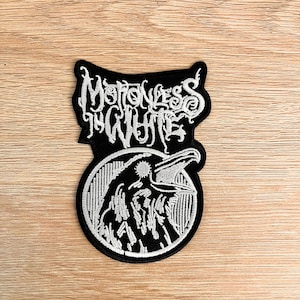 Puede incluir: Parche bordado en blanco y negro con el texto "Motionless in White" sobre un diseño circular. El diseño circular representa un cuervo con el pico abierto. El parche está sobre una superficie de madera.