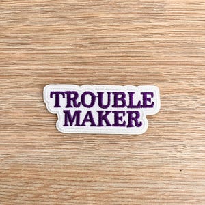 May include: White embroidered patch with purple text that reads "Trouble Maker".