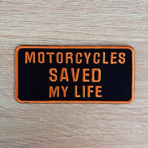 May include: Black rectangular patch with orange embroidered text reading "MOTORCYCLES SAVED MY LIFE". The patch has an orange border, designed to be sewn onto clothing or accessories. A biker-themed item.