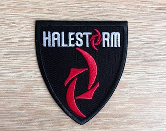 Puede incluir: Parche negro con la palabra "HALESTORM" en blanco, arqueada en la parte superior. Un diseño estilizado en rojo se encuentra debajo del texto. El parche tiene forma de escudo con borde negro.