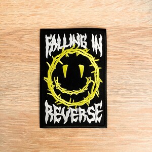 May include: A rectangular black embroidered patch featuring a yellow smiley face encircled by a barbed wire design. The text FALLING IN is embroidered in white above the smiley face, and the text REVERSE is embroidered in white below it.