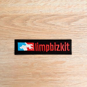 Pode incluir: Patch retangular preto com a palavra "limpbizkit" bordada em vermelho. O patch apresenta um logotipo com uma figura branca dentro de um retângulo azul e vermelho. Projetado para ser costurado em roupas ou acessórios.