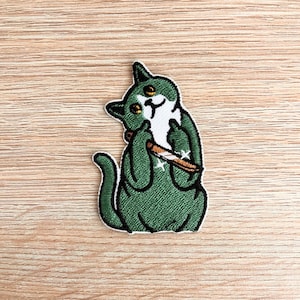 Può includere: Toppa ricamata raffigurante un gatto verde con dettagli bianchi, che tiene una sigaretta accesa. Il gatto ha occhi gialli e un'espressione birichina. La toppa è bordata di bianco ed è destinata a vestiti o accessori.