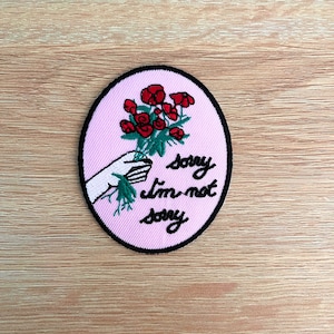 May include: Embroidered patch with a pink oval background and a black border. The patch features a hand holding a bouquet of red flowers, with the text "sorry I'm not sorry" in black script.