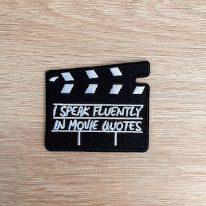 Puede incluir: Un parche negro con forma de claqueta de cine con detalles bordados en blanco. El texto "I SPEAK FLUENTLY IN MOVIE QUOTES" está bordado en blanco. El parche está sobre una superficie de madera marrón claro.
