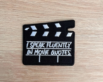 May include: A black patch in the shape of a film clapperboard with white embroidered details. The text "I SPEAK FLUENTLY IN MOVIE QUOTES" is embroidered in white. The patch is on a light brown wooden surface.