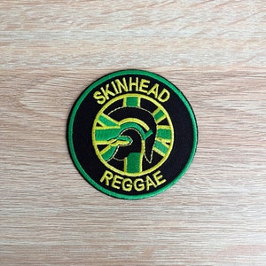 May include: A black and green circular patch with the text "SKINHEAD REGGAE" in yellow lettering. A green and yellow stylized image of a helmet is in the center of the patch.