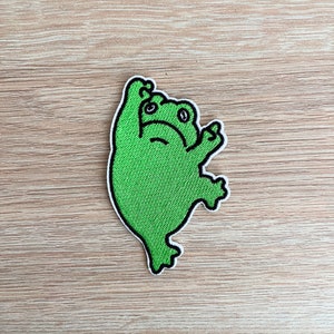 May include: Green embroidered frog patch with black outlines. The frog is hanging upside down with its arms raised and its legs spread out.