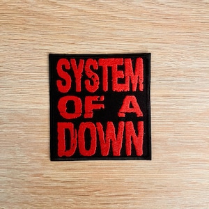 Può includere: Toppa quadrata nera con la scritta "SYSTEM OF A DOWN" ricamata in rosso. La toppa è su una superficie di legno marrone chiaro.