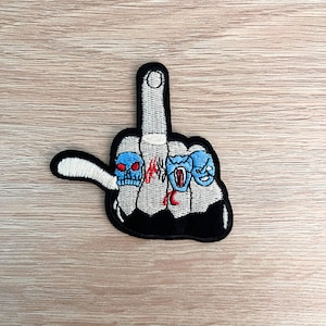 May include: A black and white embroidered patch of a middle finger with three rings on it. The rings are blue and white with red eyes and teeth. The middle finger is raised in a rude gesture.