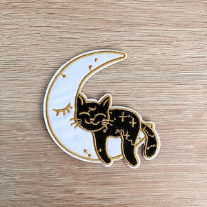 May include: Black cat sleeping on a white crescent moon with gold embroidery. The cat has closed eyes and a small smile. The moon has a gold outline and small stars.