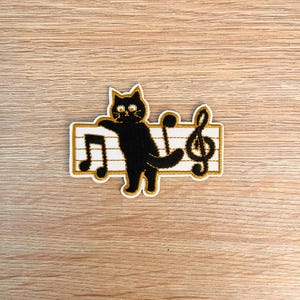 May include: An embroidered patch featuring a black cat standing on a white musical staff with gold borders. The cat has gold eyes and is positioned between a black musical note on the left and a black treble clef symbol on the right side.