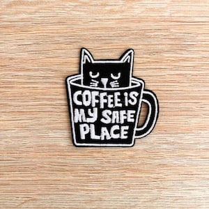 May include: Black and white embroidered patch featuring a cat inside a coffee mug. The mug has the text "COFFEE IS MY SAFE PLACE" in white. The patch is rectangular with a black border.