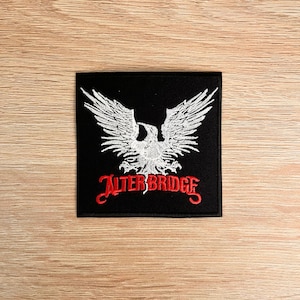 Op de afbeelding: Zwart vierkantige patch met een witte adelaar met uitgestrekte vleugels en de rode tekst "ALTER BRIDGE" eronder. De patch ligt op een lichtbruin houten oppervlak.