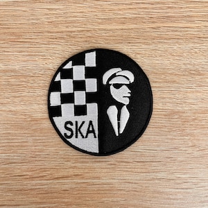 Puede incluir: Parche bordado en blanco y negro con un patrón a cuadros, una silueta de una persona con sombrero y gafas de sol, y la palabra "SKA" en texto blanco.