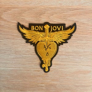 Peut inclure: Écusson brodé avec l'inscription "BON JOVI" en or. Le patch a une bordure noire et un cœur doré avec des ailes et un motif d'épée. Il est posé sur un fond en bois brun clair.