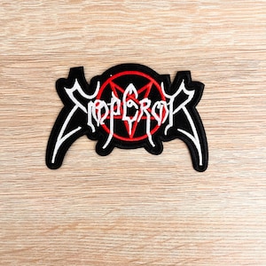 Puede incluir: Parche negro con la palabra "EMPEROR" en blanco sobre un pentagrama rojo. El parche tiene una forma única con extensiones estilizadas. Probablemente es merchandising de una banda musical.