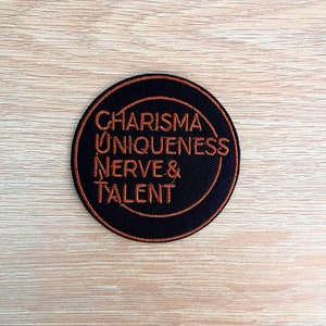 May include: A black circular patch with a white outline and the words "CHARISMA UNIQUENESS NERVE & TALENT" in orange embroidery.