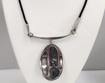 May include: A silver necklace with a black cord and an oval pendant featuring an abstract design with a small cluster of silver beads.