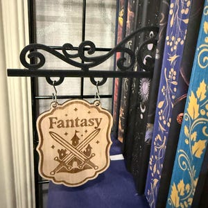 May include: A wooden sign with the word "Fantasy" and crossed swords and a castle design. The sign hangs from a black metal bracket. Books with decorative spines are in the background.