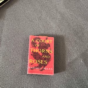 May include: A red paperback book with the title "A Court of Thorns and Roses" by Sarah J. Maas. The cover features a black illustration of a dragon.
