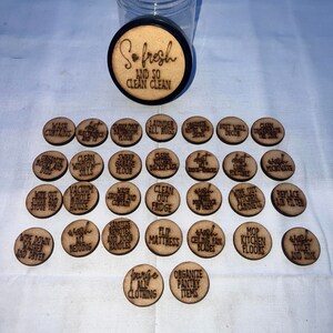 May include: A set of wooden tokens with cleaning tasks written on them. The tokens are in a clear jar with a black lid. The lid has the words "So fresh and so clean clean" written on it.