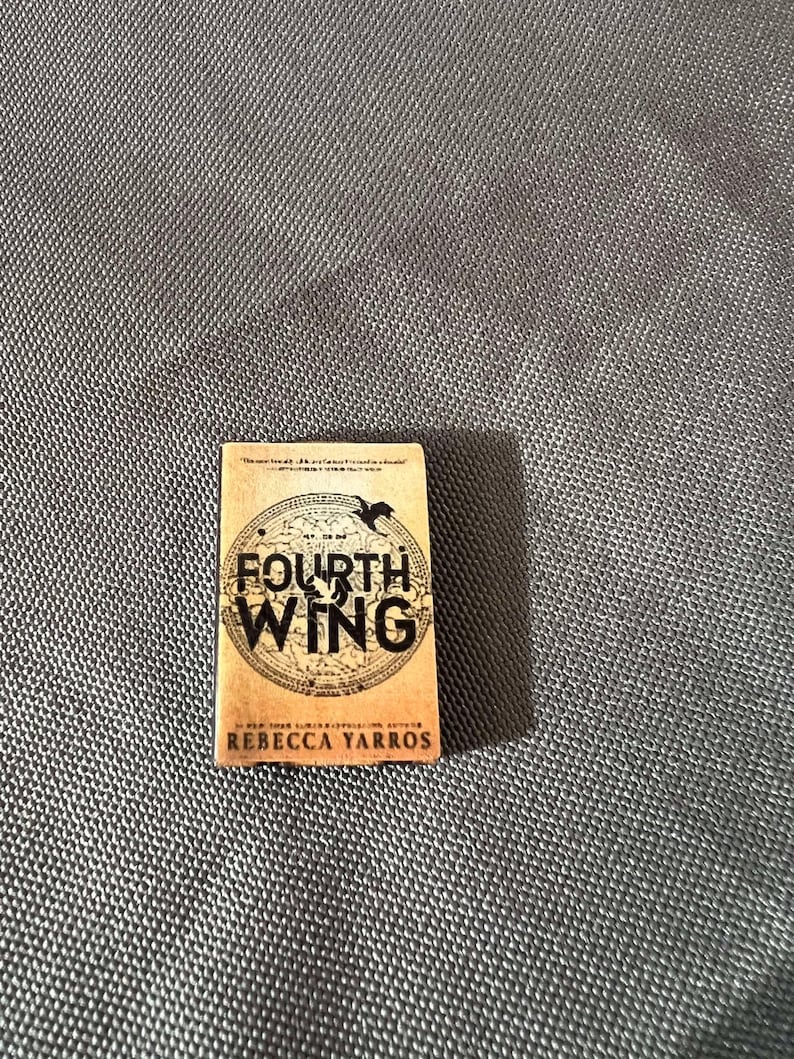 May include: A paperback book with a tan cover featuring a black and white illustration of a bird in a circle with the text "Fourth Wing" and the author's name "Rebecca Yarros".
