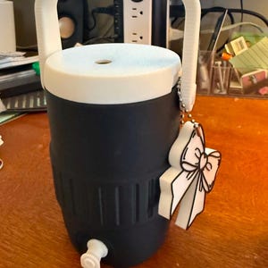May include: A navy blue and white water cooler with a white handle and a white lid. The cooler has a spigot and a decorative bow-shaped charm. The cooler is designed to keep beverages cold.